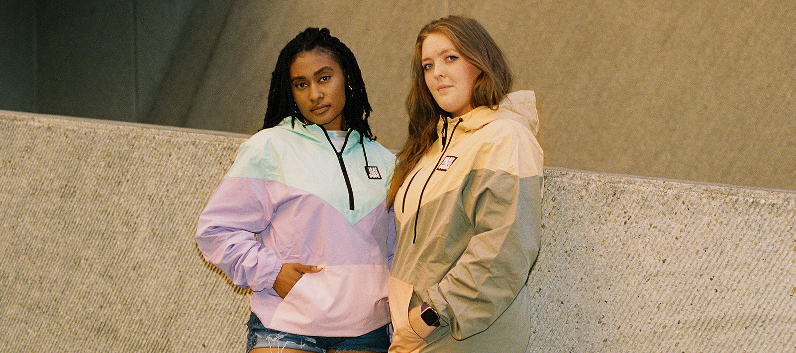 two women stand in front of a concrete half-wall, wearing neat dude windbreakers